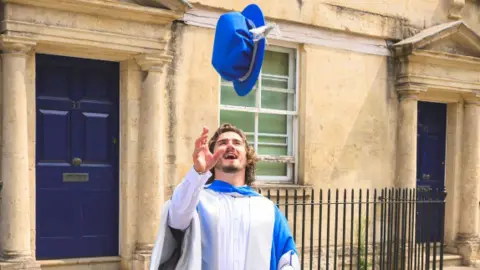 A man in a blue and white graduation gown throwing his graduation hat into the air as he smiles openly and watches his hat. His right arm is raised slightly as holds his hand open ready to catch the cap.
He is standing in front of a pavement and some Bath houses with dark doors. 