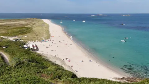 A beach on herm. The water is clear blue and the beach sandy white. The image is taken from a drone looking down onto the beach. Small dots of people can be seen relaxing on it.