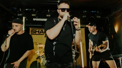 @aprilmphoto James Chakraverty (centre) performing on stage with his band Shade UK. There is a man singing into a microphone to the left and a man playing a guitar to the right. James Chakraverty is wearing a black t-shirt, dark black sunglasses and is wearing a silver necklace.