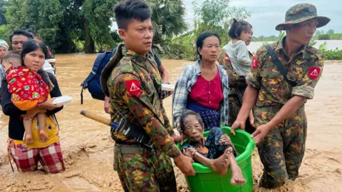 Getty Images Two soldiers carry an elderly woman in a bucket as they wade through knee deep muddy water. Behind them several other civilians, carrying children, make their way through the flooding as well.