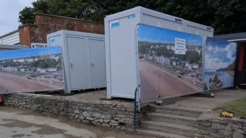 Two grey temporary toilet blocks on a raised stone platform. The blocks are surrounded by temporary barriers with an image of Filey across them.