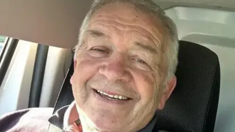 An older man smiling widely at the camera. He appears to be sitting inside a vehicle
