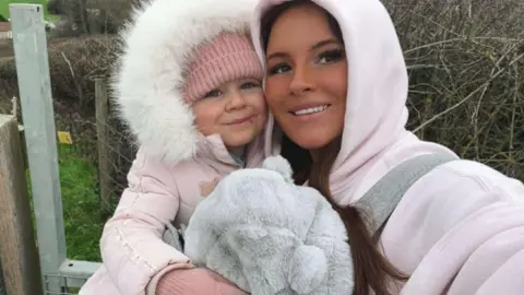 Amy-Jade Hughes An adult and a small child are standing outdoors beside a wooden gate at the edge of a field. Both are dressed warmly in pale pink winter clothing, with the adult wearing a hooded coat and carrying the child close. The child is bundled in a thick coat with a fur‑trimmed hood and is holding a large, soft grey stuffed animal.