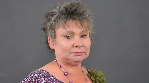 Scottish Borders Council A woman with short grey/ brown hair looking at the camera. She stands in front of a grey background. She is wearing a purple and black top and a silver and purple necklace. 