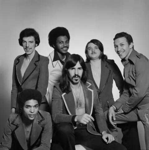Michael Putland/Getty Images Six men smile and strike a pose for a black-and-white band photo. They are wearing vintage-style jackets with wide lapels and have 1970s-style haircuts including "Afros" and long bangs.