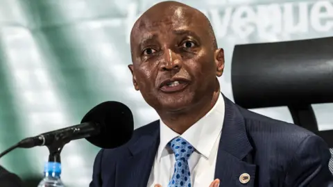 Patrice Motsepe wearing a dark blue suit with a patterned blue tie, speaks into a microphone during a press conference