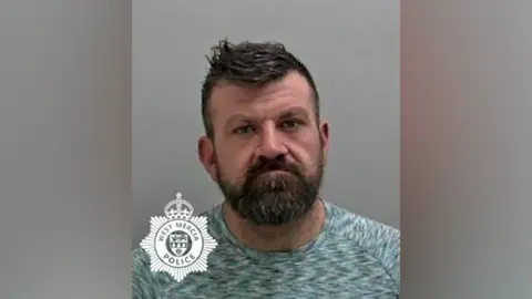 Police mugshot of Craig Nunn. He has a neutral expression on his face, a full brown beard and spikey brown hair. He is wearing a grey, crew neck t-shirt