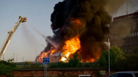 A large fire engulfs a building, with significant flames rising from the ground, creating a large black cloud of smoke, while a jet of water from an aerial hose works to fight the fire.