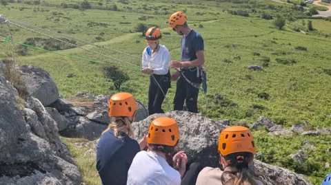 Three girls in orange helmets watch their friend abseil down a rock face with the help of an outdoor instructor.