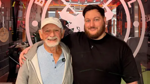 A tall man with dark hair and a beard stands with his arm around an older man with a white beard and moustache, wearing a cap