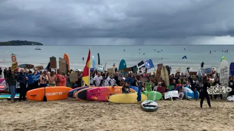 People with paddleboards and placards on a sandy beach. Behind them is the sea and grey clouds. 