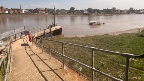 A concrete sloped platform leads down to the Great Ouse river. There is a handrail and a life preserver. The ferry is moored in the water in the distance.