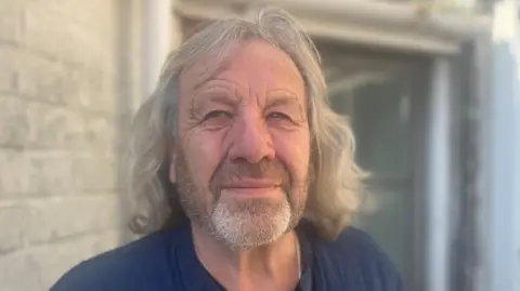 An older man looking at the camera, with grey, mid length hair and a grey beard. He is wearing a navy shirt and is not smiling. He is standing outside a house with an open door.