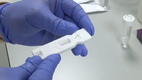 A lateral flow test for black fungus disease being held in a person's hands in rubber gloves. It is a small plastic strip with a window in it with two horizontal lines visible. 