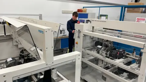 A man with dark brown hair and dark blue overalls stands in an industrial looking room surrounded by machinery. He is looking at a large grey structure with a turbine at the centre.