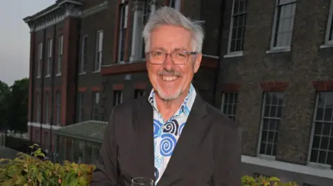 Getty Images Griff Rhys Jones smiling at the camera holding a glass. He has short silver hair, and is wearing glasses, a white shirt with blue swirls on it, and a black suit jacket. There is an imposing grey brick building behind him.