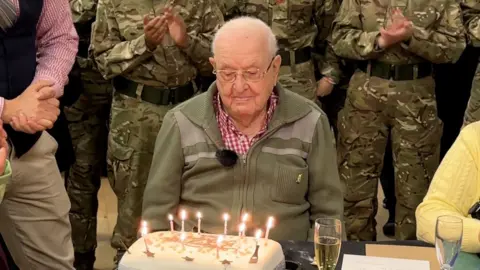 Jack Waterfield sitting at a table with a big birthday cake in front of him, with candles lit. The camouflaged uniforms of cadets can be seen behind him and they are clapping.
