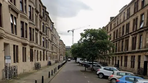 A long street of blonde sandstone tenements with some cars parked in front of them.