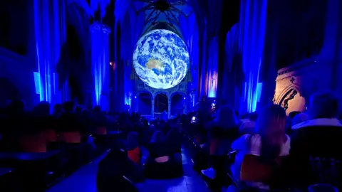 Steve Mellen/BBC People watch a musical display under the Luke Jerram artwork Gaia inside Bristol Cathedral. The internal columns of the cathedral are illuminated by blue light and Gaia, a to-scale model of the earth, is illuminated