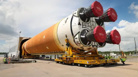 NASA Nasa's SLS rocket lying horizontally and on supports and being wheeled into an enclosure. The rocket appears gigantic in compariosn to engineers sitting alongside the vehicle.