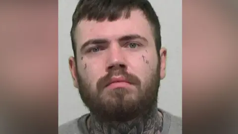 Mugshot of Byron Stubbs. He has short dark hair and a thick black beard, with small illegible symbols tattooed on his cheeks and a giant floral tattoo covering the entirety of his neck and throat