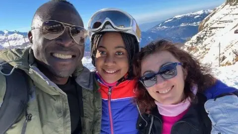 Family handout Daisy in ski gear on a mountain along with her dad (left) and mum (right). They all wear sunglasses and winter coats, and are smiling at the camera.