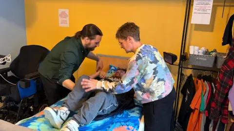 Isaac is being lowered into bed by his mother and brother. To the right is a hanging rail of t-shirts. Isaac's brother is standing on the left of the bed and is wearing a green top. His mum is wearing a flowery top. Both have their arms in the air mid-lift.