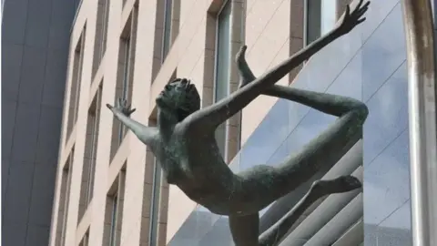 BBC A naked girl made out of metal with a green tinge has her arms outstretched and her legs thrown back behind her. She appears to be floating.
