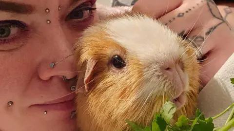 A close-up showing part of a woman's face, snuggling up to a guinea pic. The woman has several facial piercings. he animal is ginger and white and has dark eyes and a pink nose. It is very cute