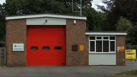 Cambridgeshire Fire and Rescue An outside view of Gamlingay fire station. It is a brick building with a large red door in the middle. Attached on the right is a lower building with a large window. Behind it are trees.
