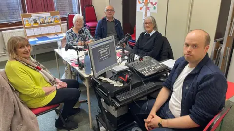 Three women and two men sit around a table with three microphones on it and a mixing desk. The woman on the left is wearing a long silk scarf, a lime jumper and black trousers. The woman at the head of the table is wearing a patterned blouse of blue, purple and green colours. A man sits beside her wearing glasses, a navy jacket, grey jumper and a checked shirt. The lady right of him wears glasses, a black jumper with a light collar visible. The man sat at the end of the table wears a white t-shirt with an open navy shirt and jeans. There is a window at the back of the room behind a noticeboard with photographs of puppies on it. Cabinets and stacks of chairs can also be seen in the background.