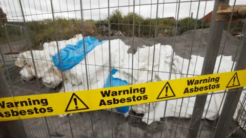 PA Media A pile of plastic sheets rolled up on the ground, with a metal fence and yellow tape reading "warning asbestos" in front of it.