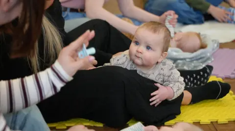 A baby of around 6 months old with fair hair and blue eyes pushes itself up on its mother's leg who is sitting on the floor at a mother and baby group, though she is not fully in shot. The baby is wearing a beige romper suit with a dinosaur pattern. It is looking towards the hand of a woman sitting next to it, also not in shot. She is holding a blue dummy and her stripy cream and maroon sleeve is visible. There are yellow rubber mats on the floor and another baby with red hair can be seen in the background lying on its back on a grey blanket and holding its mum's hands.