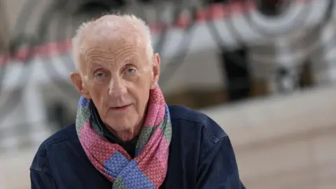 Getty Images Paul Costelloe has receding grey hair. He is wearing a bright pink, blue and green scarf and a blue jacket.