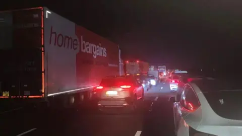 Traffic at a standstill on a dark three-lane road. There is a mix of cars and lorries, lots with their red brake lights on. 