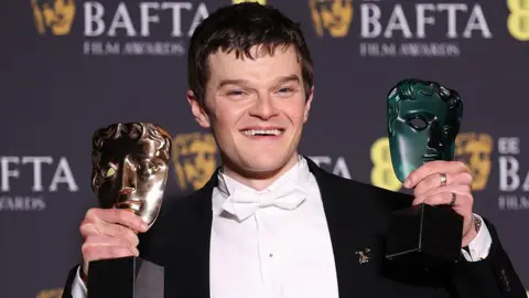 Getty Images Head and shoulders shot of Robert Aramayo celebrating at the Baftas. He is wearing a white shirt and bow tie and a black jacket. He is holding up his awards and smiling.