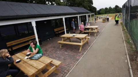 Oxford City Council People sitting at or standing around outdoor tables at a café opposite a tennis court. It is an overcast day.