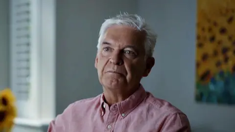  Burning Bright Productions/PA Phillip Schofield looking sombre, in a pink shirt in a room with sunflowers plus a sunflower picture, filming for episode two of Channel 5's Cast Away.