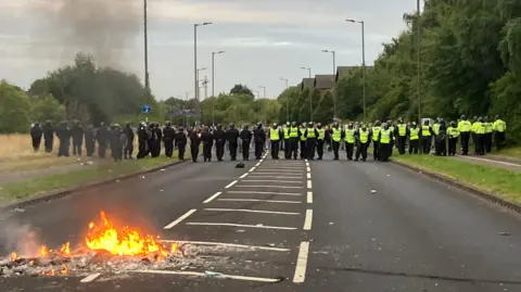 A fire burns in the middle of a road. Behind it, a line of dozens of police officers stretches from one side of the road to the other. Half of them are wearing hi-vis vests over their uniforms.