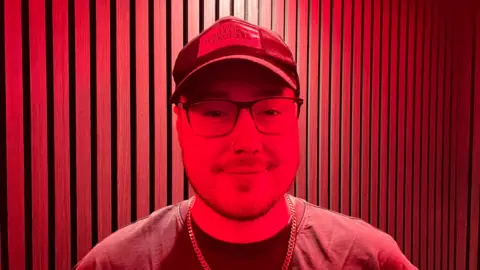 Evo standing in a recording studio with red lighting. He has a beard and nose ring, is wearing a black North Face cap, glasses, black t-shirt and gold chain.
