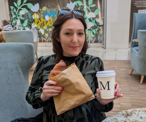 Hayley Turner Jo is sitting in a cafe. She is wearing a green and black dress and holding a croissant and a cup of coffee. She has brown, shoulder length hair, and is wearing sunglasses on her head. 