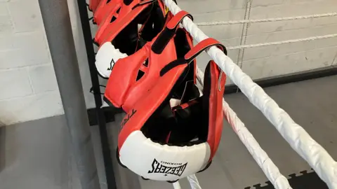 Three white and red boxing gloves hung up on the white ropes surrounding a boxing ring. The gloves are lined up and at the centre of the image. There is a white wall behind and the floor is grey.