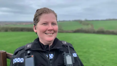 A policewoman in uniform stands with fields behind her in the countryside