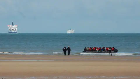 BERNARD BARRON/AFP via Getty Images Police officers on a sandy beach in France walk in the direction of an inflatable boat carrying migrants. In the distance are two ferries. 
