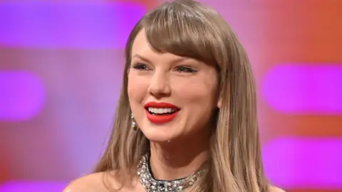 PA Media Taylor Swift smiling, wearing her long, blonde hair down to her shoulders, with dark eyeliner and red lipstick, and a dress with bright studs on the neckline, against the purple and orange background of the Graham Norton Show