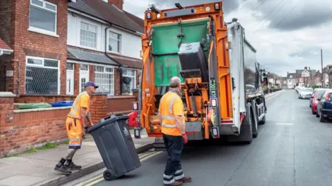 A stock image showing a bin lorry and two workers, both wearing orange hi-vis, with a wheelie bin