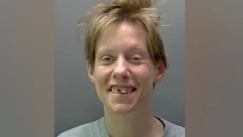 Hertfordshire Police Erin McNeilly is seen in a police custody image the woman with short brown hair, smiling. She is wearing a grey round-necked T-shirt and one of her front teeth is missing.