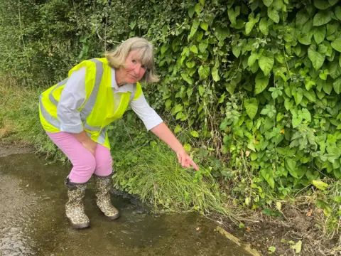 Sue Wooley - a woman with bobbed hair - is pointing to a stream of water on the ground. She is wearing leopard print wellington boots, pink jeans, a white shirt and a yellow high-vis jacket. The water on the ground is underneath her feet and is leaving mud on the path.