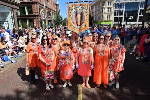 A group of women in orange dresses and sashes wearing sunglasses as they pose on a street in Belfast and hundreds of spectators are behind them. The women are holding a banner that says "Sister Clarke Memorial L.L.O.L No. 156 Parkhead Dist 2". The sun is shining on the women. 