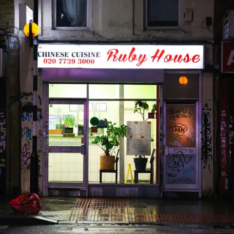 Harriet Armstrong Ruby House Chhinese takeaway with plants on chairs in the window beside a door covered in graffiti
